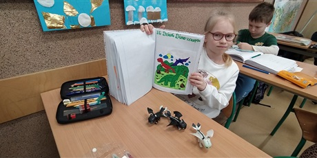 Powiększ grafikę: dziewczynka siedzi przy stoliku i trzyma zeszyt z kolorowanką dinozaurów