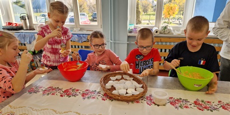 Powiększ grafikę: dzieci siedzą przy stole trzymają miski i wyrabiają w nich ciasto na babeczki i wkładają do foremek