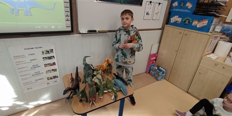 Powiększ grafikę: przy stoliku z figurkami dinozaurów  stoi chłopiec i opowiada o nich