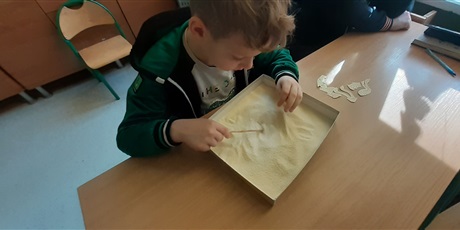 Powiększ grafikę: Zabawa w paleontologa - chłopiec z 1c szuka elementów papierowego szkieletu dinozaura w kaszy mannej, imitującej piasek.