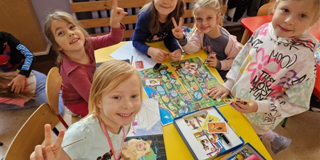Powiększ grafikę: W sali przy stoliku siedzą uśmiechnięte dziewczynki,grają w grę planszową.  