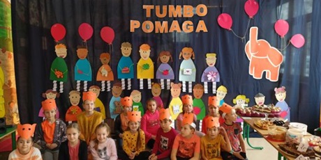 Powiększ grafikę: Grupa dzieci siedzi na tle dekoracji z napisem Tumbo pomaga.