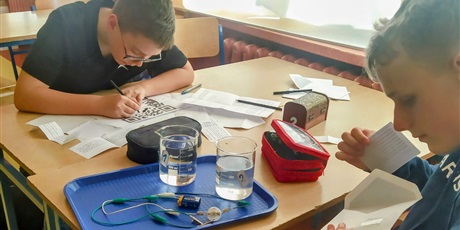 Powiększ grafikę: Dwoje chłopców siedzą przy stoliku, jeden koloruje kod QR, drugi wyjmuje zagadkę z koperty