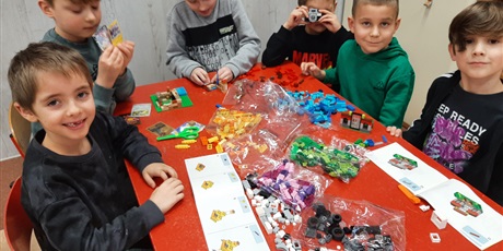 Powiększ grafikę: Grupa dzieci siedzi przy stole, budując z klocków Lego wg instrukcji.