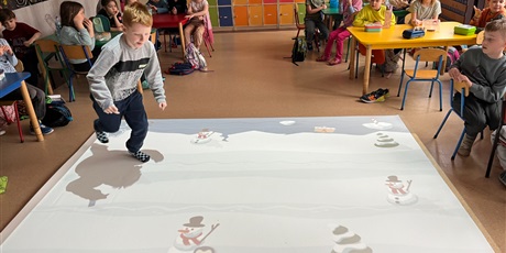 Powiększ grafikę: Chłopiec próbuje złapać pingwina na interaktywnym dywanie, pozostałe dzieci siedzą przy stolikach przyglądają się.