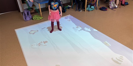 Powiększ grafikę: Dziewczynka próbuje złapać pingwina na interaktywnym dywanie, pozostałe dzieci siedzą przy stolikach przyglądają się.