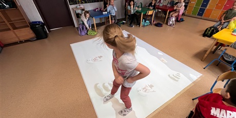 Powiększ grafikę: Dziewczynka próbuje złapać pingwina na interaktywnym dywanie, pozostałe dzieci siedzą przy stolikach przyglądają się.