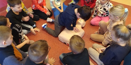 Powiększ grafikę: Chłopiec klęczy na podłodze, uciska klatkę piersiową fantoma, wokół siedzą dzieci i patrzą.