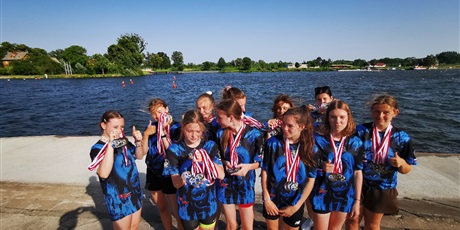 Powiększ grafikę: Na tle akwenu wodnego stoi 11 dziewcząt w strojach sportowych, w rękach lub na szyi mają medale.