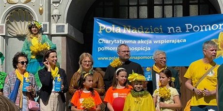 Powiększ grafikę: na tle Bramy Artusa i niebieskiego plakatu stoją dorośli i dzieci ubrani na żółto