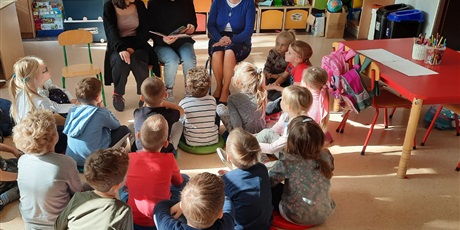 Powiększ grafikę: Grupa dzieci siedzi na podłodze i słucha czytanych tekstów.
