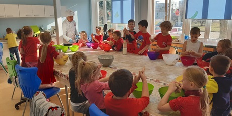 Powiększ grafikę: Dzieci siedzą na krzesłach dookoła stołu i w miskach mieszają kolejno dodawane składniki wg zaleceń Pana kucharza. Pan kucharz stoi przy stole.