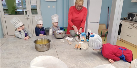 Powiększ grafikę: Dzieci w kucharskich czapkach patrzą, jak pani przygotowuje różne produkty do ciasta.