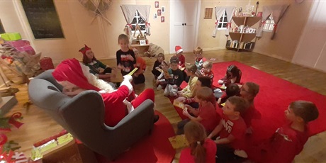 Powiększ grafikę: Dzieci z klasy 1c siedzą w pokoju Mikołaja na czerwonym dywanie. Dziewczynka odbiera dyplom z rąk Mikołaja.