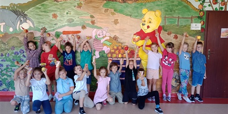 Powiększ grafikę: Dzieci stoją na tle jesiennego ogrodu Kubusia Puchatka, są uśmiechnięte,  trzymają w rękach jabłka i soki jabłkowe.