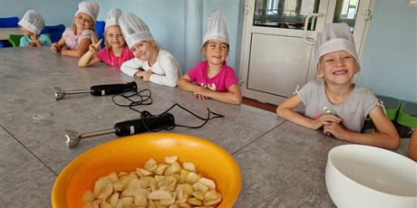 Powiększ grafikę: Dziewczynki siedzą przy stole,są uśmiechnięte,przed nimi stoi wielka miska pełna pokrojonych jabłek.