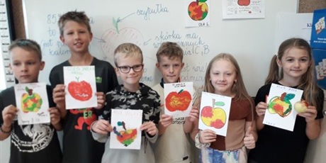 Powiększ grafikę: Uczniowie stoją pod tablicą i pokazują wykonane prace plastyczne z plasteliny, na których są wyklejone jabłka