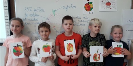 Powiększ grafikę: Uczniowie stoją pod tablicą i pokazują wykonane prace plastyczne z plasteliny, na których są wyklejone jabłka