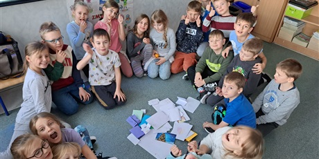 Powiększ grafikę: Grupa dzieci siedzi w kręgu, a w środku kartki z pozdrowieniami.
