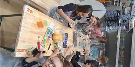 Powiększ grafikę: Grupa dzieci siedzi przy stole i maluje farbami gipsowe doniczki.