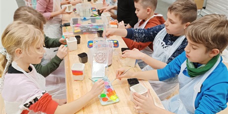 Powiększ grafikę: Grupa dzieci siedzi przy stole i maluje farbami gipsowe doniczki.