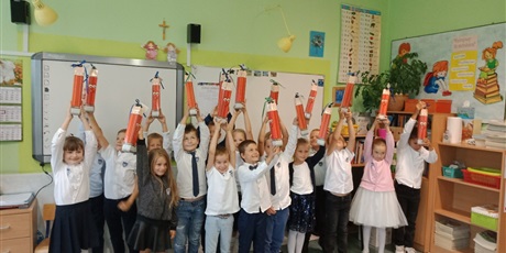 Powiększ grafikę: Grupa dzieci w strojach galowych podnosi do góry ręce, w których trzymają pudełka w kształcie ołówka.