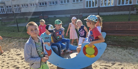 Powiększ grafikę: dzieci stoją na placu zabaw i bujają się na niebieskiej huśtawce