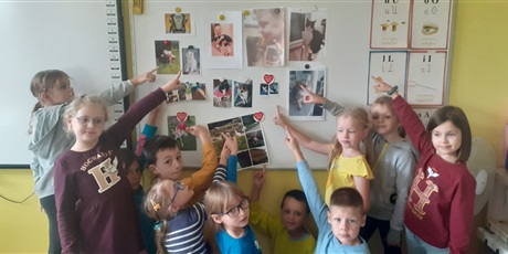 Powiększ grafikę: Dzieci stoją przy tablicy i pokazują zdjęcia swoich domowych zwierząt.