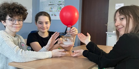 Powiększ grafikę: Uczennice trzymają kolbę z płynną zawartością i naciągniętym na kolbę czerwonym balonikiem. Balonik zwiększa objętość
