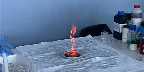 Powiększ grafikę: Na stole demonstracyjnym, na porcelanowej podstawce widać szary pył i powstający pomarańczowy ogień. Z boku- odczynniki chemiczne i szkło laboratoryjne