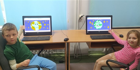 Powiększ grafikę: dwoje dzieci z 1b siedzi przy komputerach i pokazują planetę Ziemię wykonaną w programie Paint